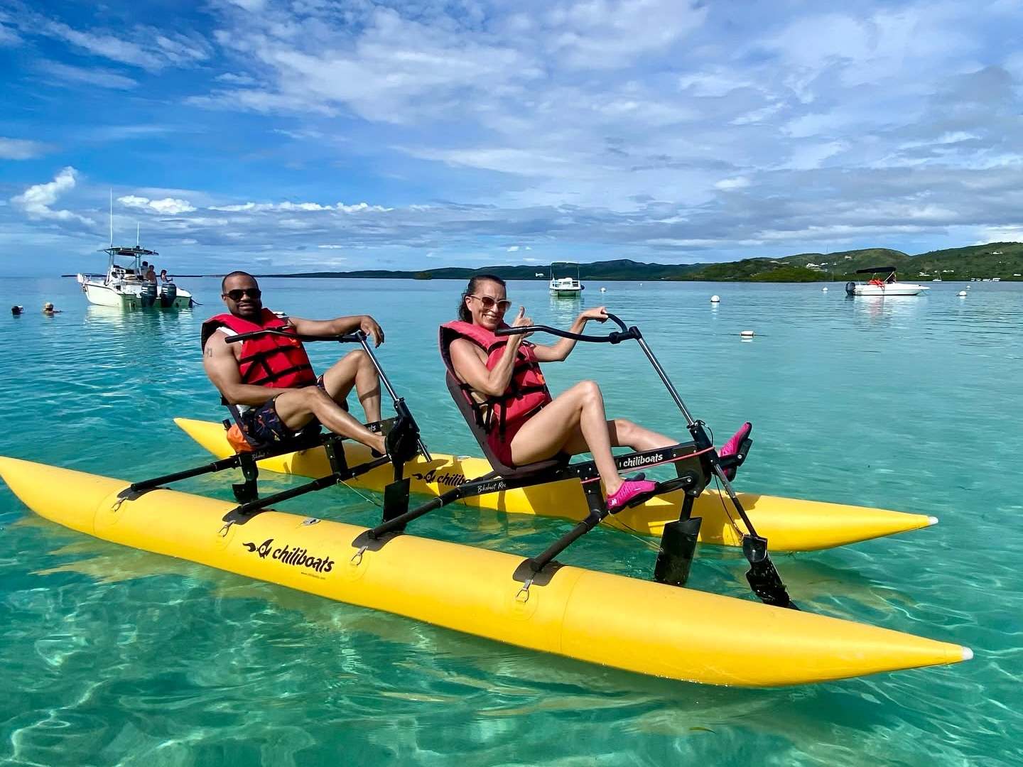 Two people on a yellow water bike in clear blue sea under a cloudy sky.