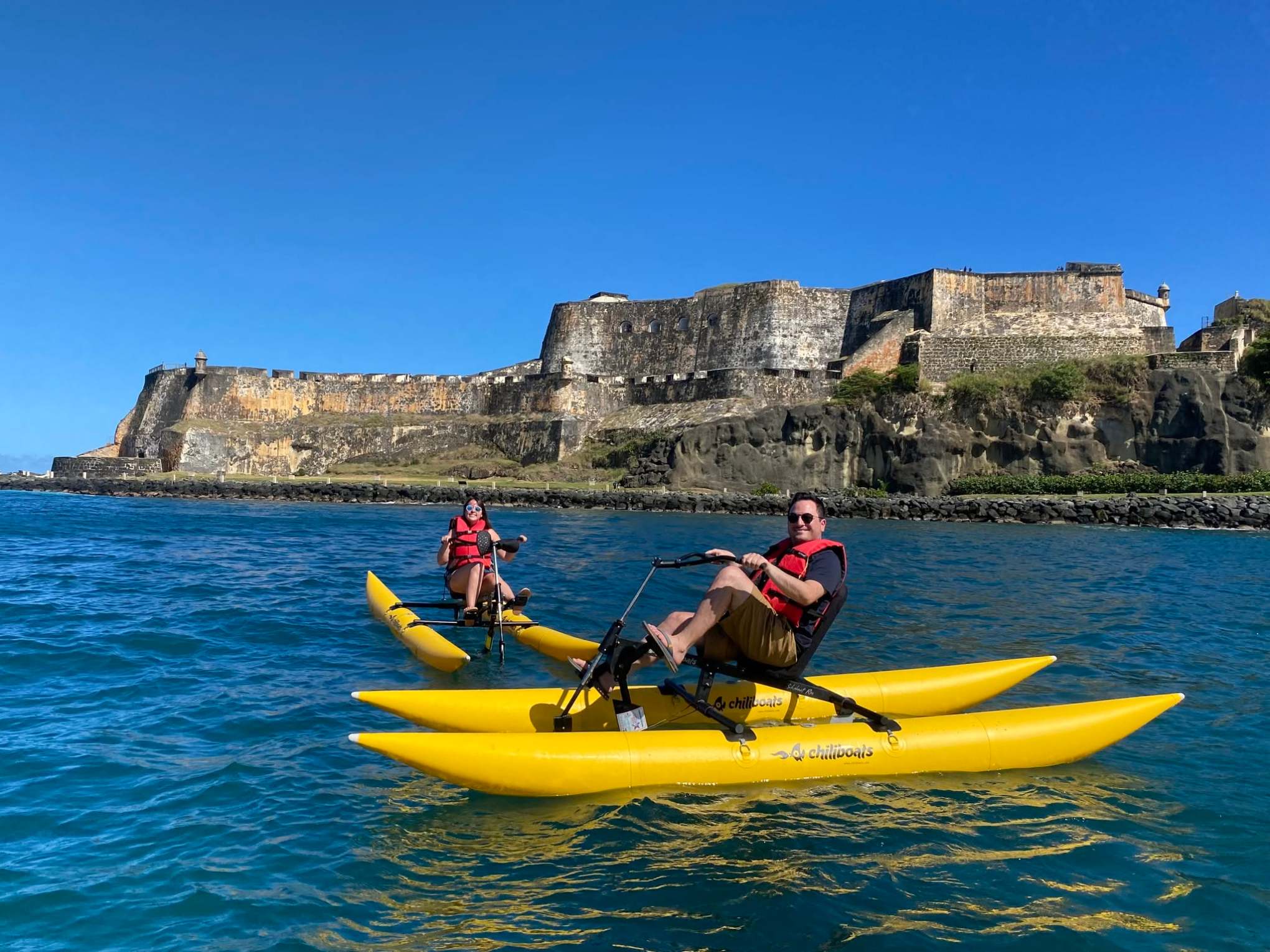 Two people pedal yellow water bikes near a historic fort on a sunny day.