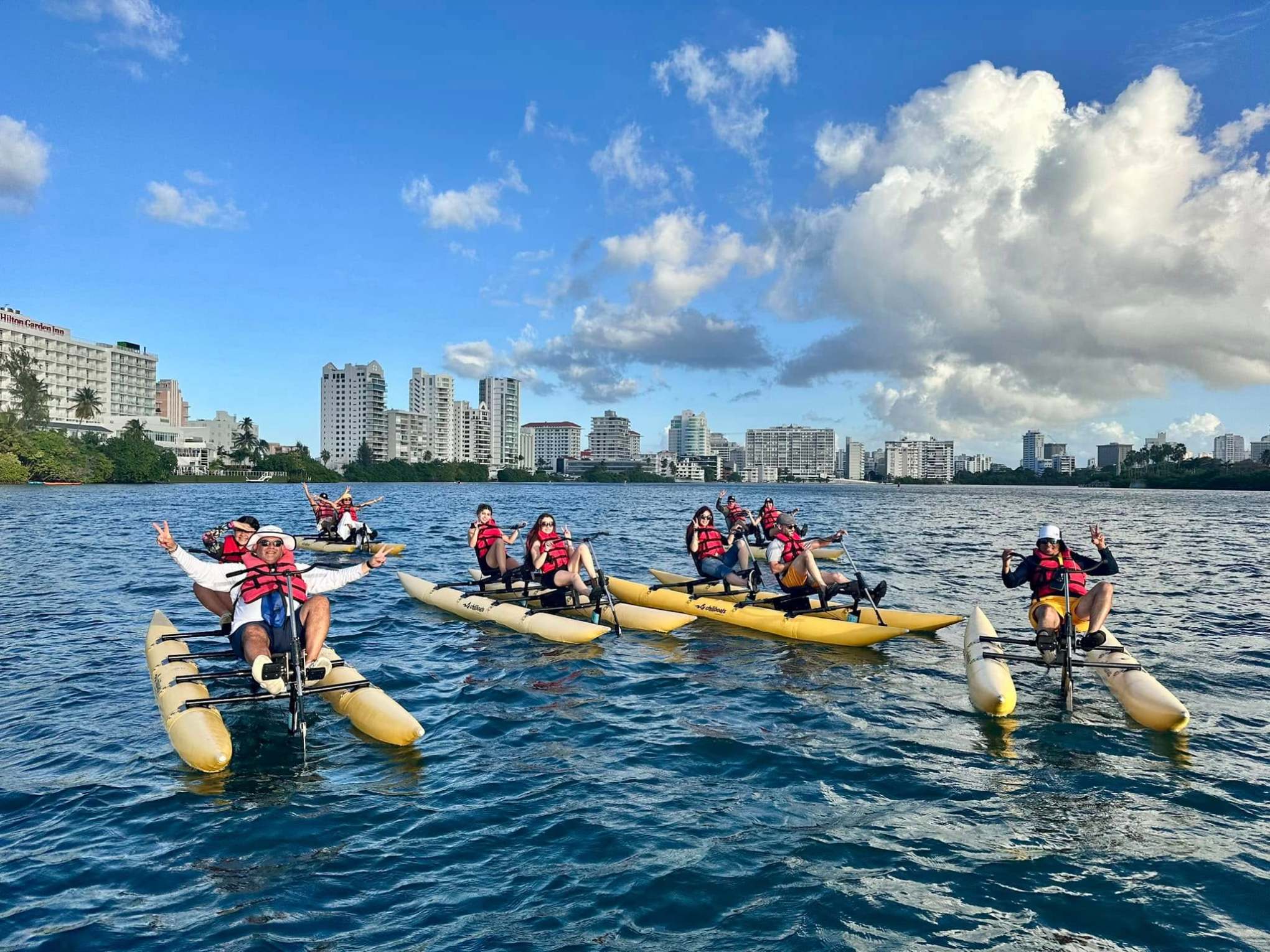 Group kayaking on water with city skyline in background, under a partly cloudy blue sky.