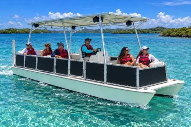 Pontoon boat with six people wearing life jackets on clear blue water.