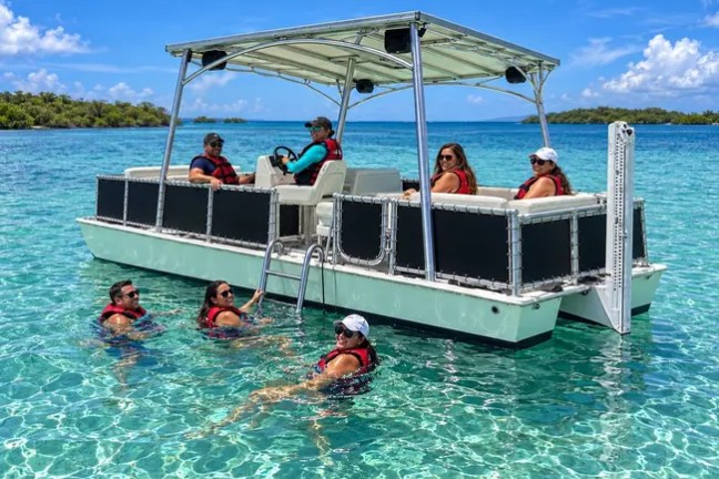 People on a pontoon boat float in clear blue water, some swimming nearby.