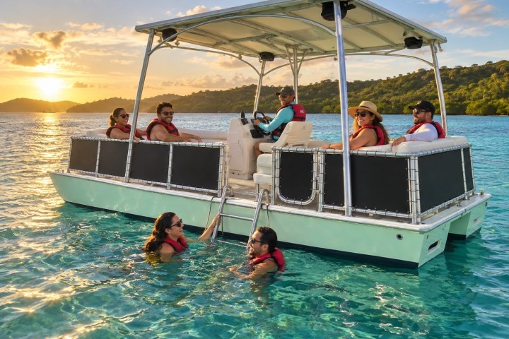 People on a pontoon boat in clear water during sunset, two swimmers nearby.